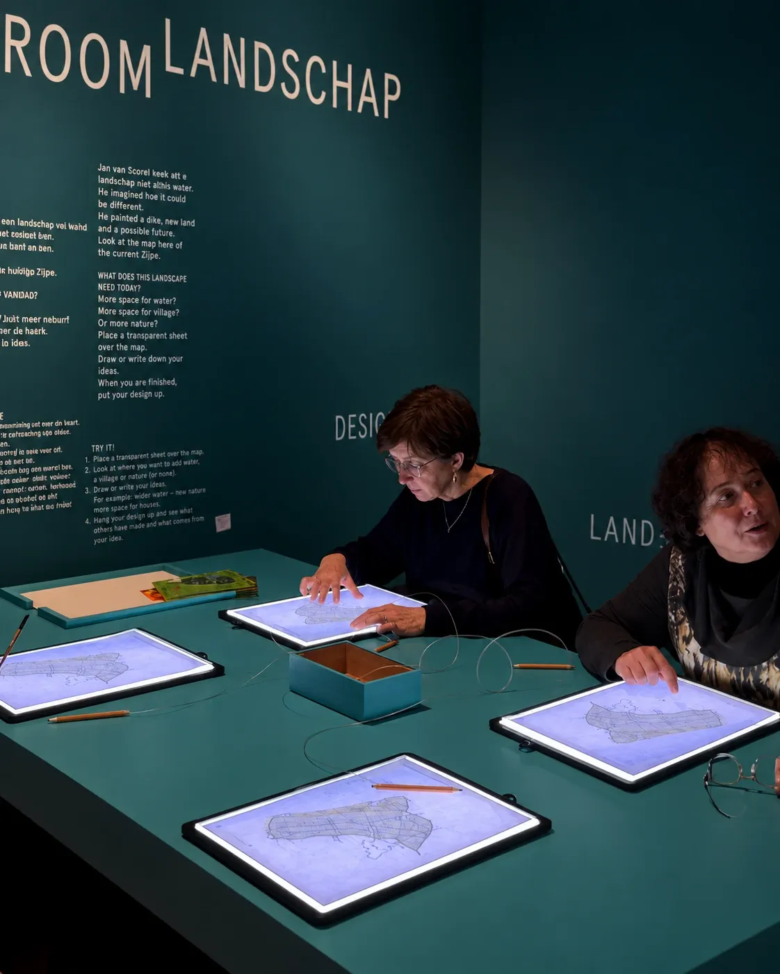Enhanced video still of visitors drawing landscapes on the illuminated maps at the teal museum table, beneath the Dutch exhibition instructions.