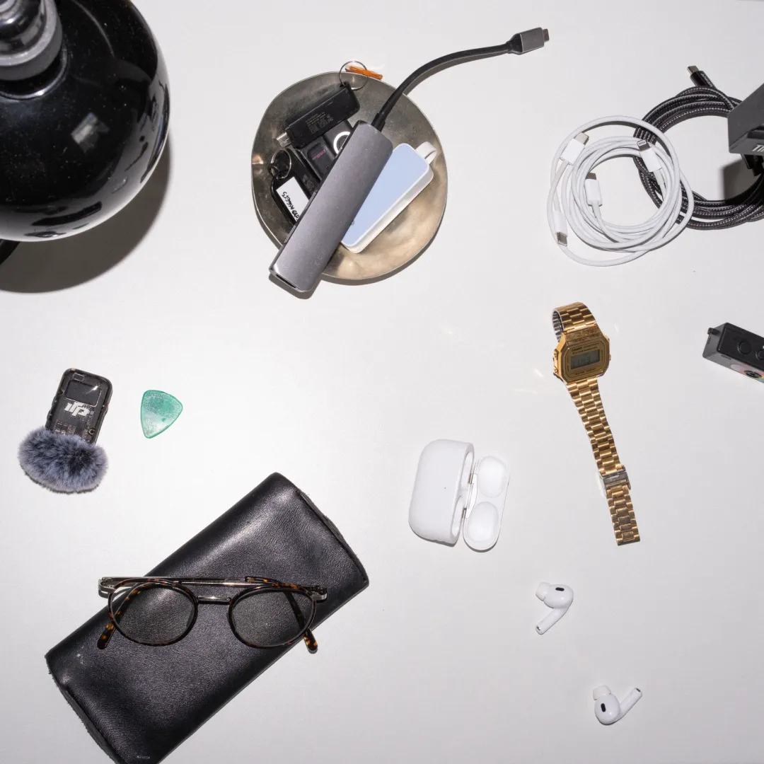 Flat-lay of James's white desk — glasses, a wireless microphone, a gold watch, earbuds and cables