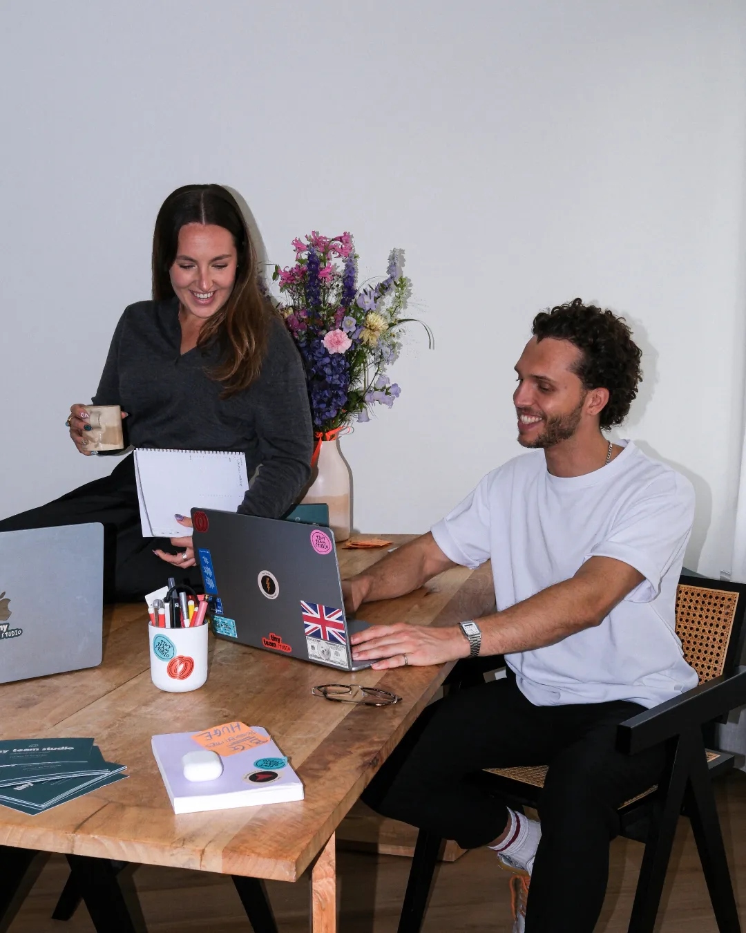 Katie and James laughing together at a wooden table — she holds a notebook and coffee, he types on a sticker-covered laptop